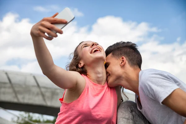 Молода пара дає поцілунок сидячи в місті на відкритому повітрі з телефоном, приймаючи селфі — стокове фото