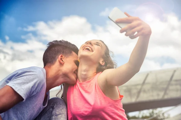 Pareja joven dando beso sentado en la ciudad al aire libre con teléfono tomando selfie — Foto de Stock
