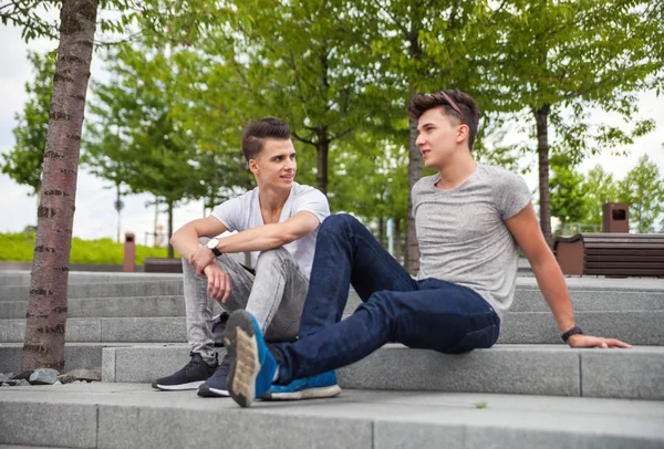 Dos amigos ropa casual sentado en las escaleras de la ciudad y hablando, b —  Fotos de Stock