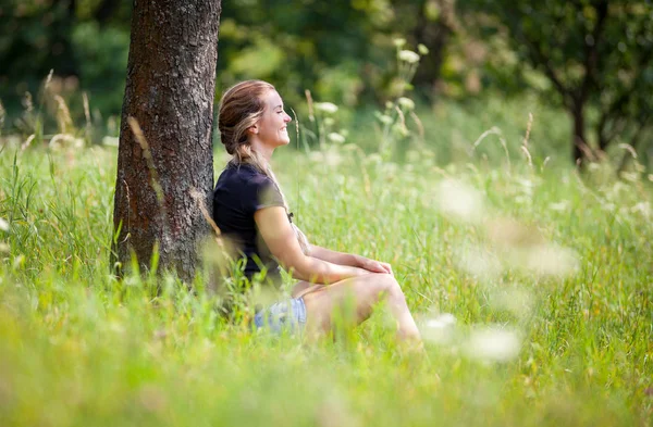 Ontspannen meisje zittend onder de boom op weide op zomerdag — Stockfoto