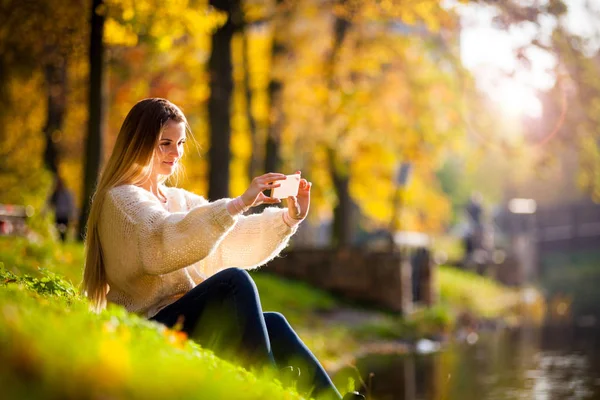 Mujer por lago en colorido parque de otoño tomando fotos con teléfono móvil — Foto de Stock