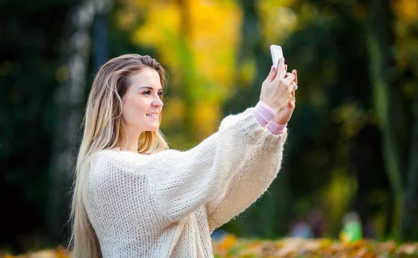 Woman in warm sweater taking picture by mobile phone walking in autumn park — Stock Photo, Image