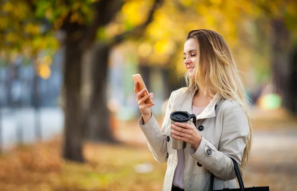 Красива жінка використовує смартфон, що йде барвистою осінньою вулицею — стокове фото