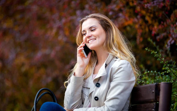 Happy woman talking smartphone sitting on bench at colorful autumn street — Stock Photo, Image