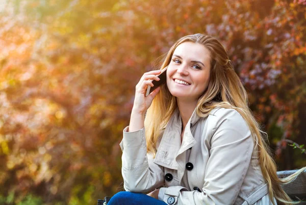 Gelukkige vrouw met behulp van de smartphone zit op bankje aan kleurrijke herfst straat — Stockfoto