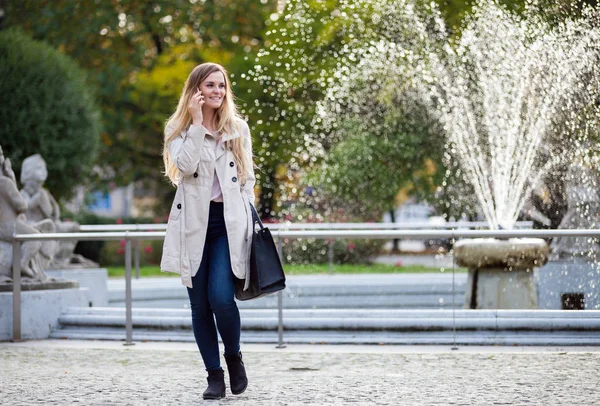 Улыбающаяся женщина в куртке гуляет по городу и разговаривает по смартфону — стоковое фото