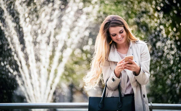 Mujer sonriente vistiendo abrigo caminando en la ciudad y usando smartphone —  Fotos de Stock