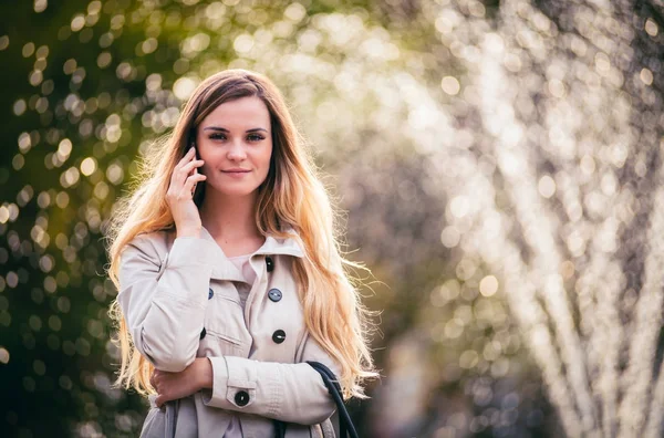 Smiling woman wearing coat walking in city and using smartphone — Stock Photo, Image