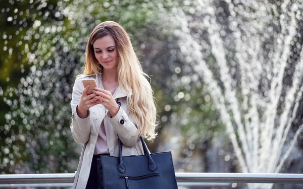 Usmívající se žena nosí kabát procházky v městě a pomocí smartphonu — Stock fotografie