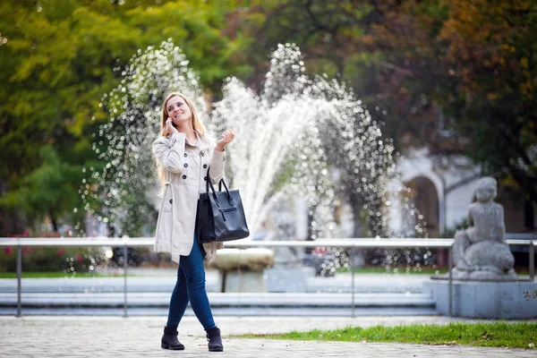 Femme souriante portant un manteau marchant en ville et utilisant un smartphone — Photo