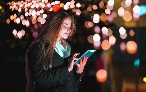 Dívka ve městě v noci pomocí tabletu sedí na lavičce — Stock fotografie