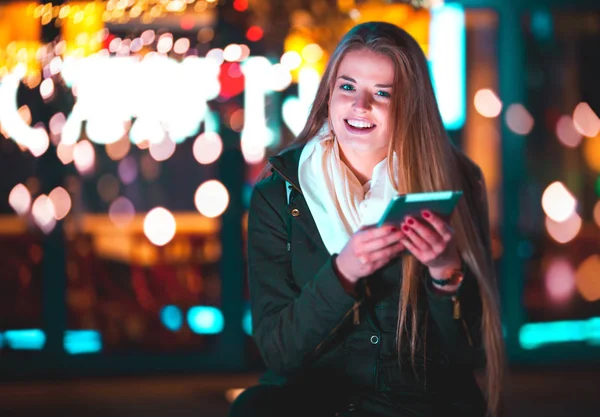 Chica en la ciudad por la noche usando tableta sentada en el banco —  Fotos de Stock