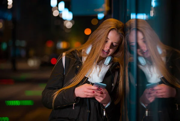 Mulher com fones de ouvido usando tablet esperando na parada de ônibus à noite na cidade — Fotografia de Stock