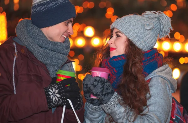 Liebespaar in warmer Kleidung mit heißen Getränken genießt Weihnachtsmarkt am Abend — Stockfoto
