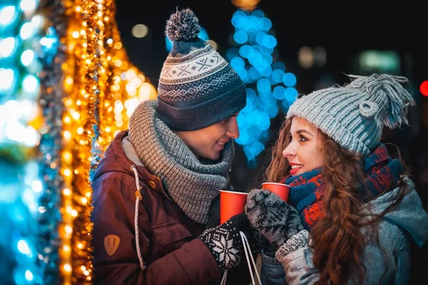 Fröhliches Paar mit Heißgetränken auf Weihnachtsbeleuchtung Hintergrund beim Abendspaziergang in der Stadt — Stockfoto