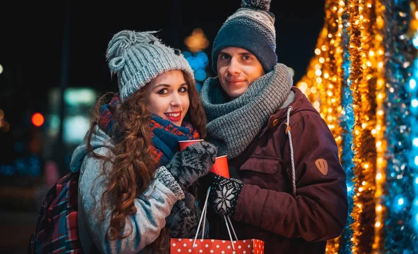 Fröhliches Paar mit Heißgetränken auf Weihnachtsbeleuchtung Hintergrund beim Abendspaziergang in der Stadt — Stockfoto