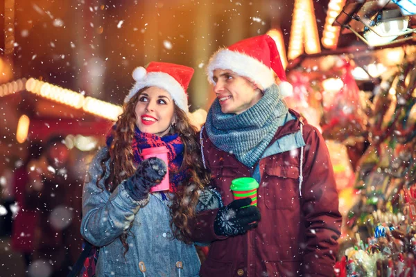 Para w ciepłe ubrania z gorącymi napojami, ciesząc się Jarmark bożonarodzeniowy z śniegu na wieczór — Zdjęcie stockowe