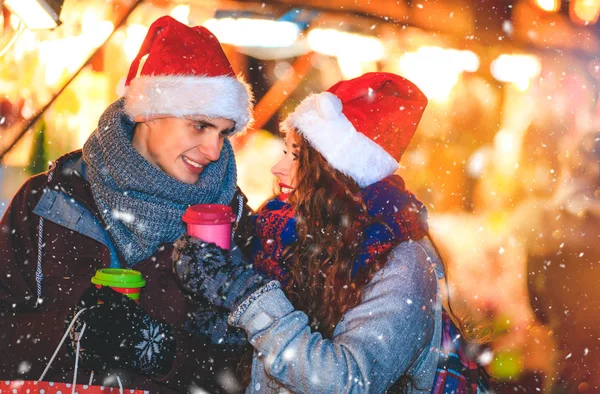 Casal em roupas quentes com bebidas quentes apreciando o mercado de Natal com neve à noite — Fotografia de Stock