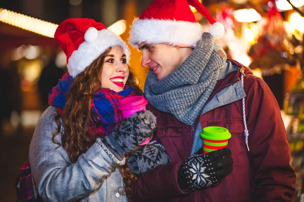 Casal feliz em roupas quentes com bebidas quentes apreciando o mercado de Natal à noite — Fotografia de Stock