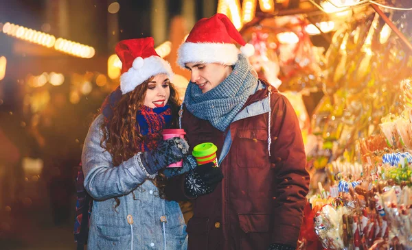 Casal feliz em roupas quentes com bebidas quentes apreciando o mercado de Natal à noite — Fotografia de Stock