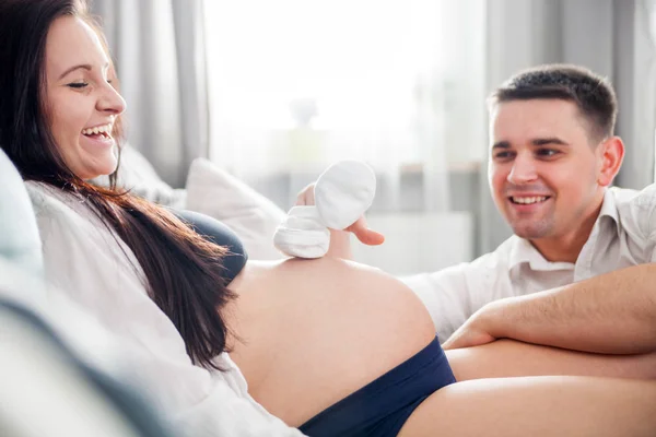 Zwangere paar spelen samen met babyslofjes zittend op de Bank thuis — Stockfoto