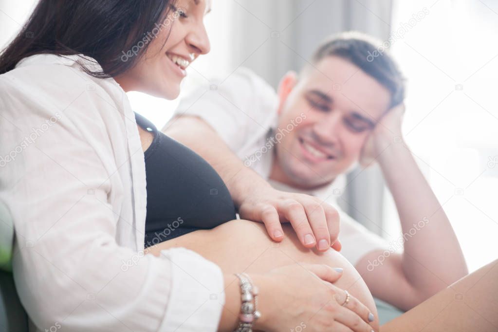 Young pregnant couple relaxing on sofa at home, touching belly