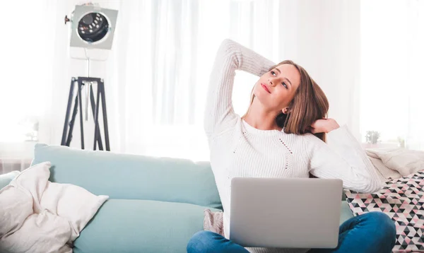 Mujer en casa sentada en el sofá y estirándose mientras trabaja con el ordenador portátil —  Fotos de Stock