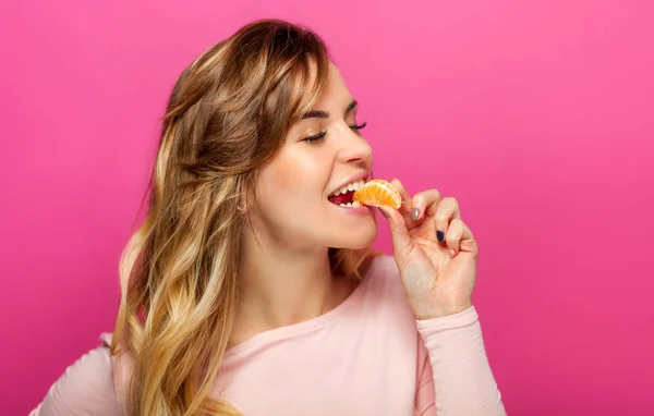 Beautiful woman on pink background eating orange fruit — Stock Photo, Image