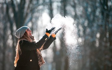 Smiling young woman throwing snow in the air at sunny winter day clipart