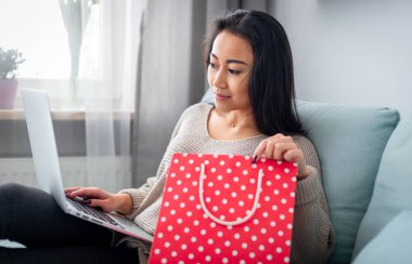 Asyalı bir kadın elinde kırmızı bir hediye çantası tutuyor ve evde dizüstü bilgisayar kullanıyor.