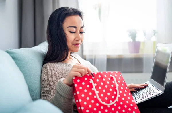 Mujer asiática sosteniendo bolsa de regalo roja y compras en línea utilizando el ordenador portátil en casa —  Fotos de Stock