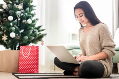 Mutlu Asyalı kız internette dizüstü bilgisayarla alışveriş yapıyor süslü Noel ağacında oturuyor.