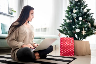 Mutlu Asyalı kız internette dizüstü bilgisayarla alışveriş yapıyor süslü Noel ağacında oturuyor.