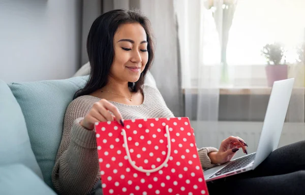 Mujer asiática sosteniendo bolsa de regalo roja y compras en línea utilizando el ordenador portátil en casa —  Fotos de Stock