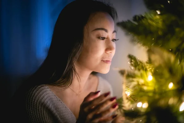 Beautiful Asian woman decorating Christmas tree in home at evening — ストック写真