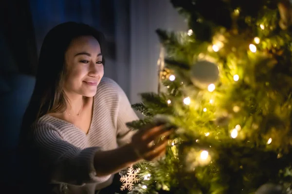 Beautiful Asian woman decorating Christmas tree in home at evening — ストック写真