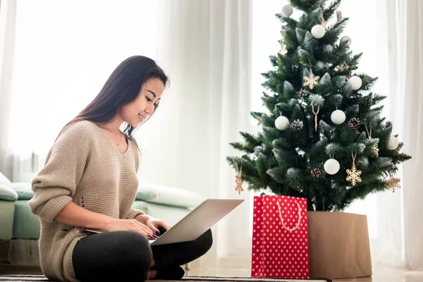 Gelukkig aziatisch meisje winkelen online met laptop zitten bij versierde kerstboom achtergrond — Stockfoto