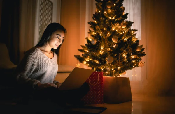 Gelukkig aziatisch meisje winkelen online met laptop zitten in de buurt versierde kerstboom achtergrond 's avonds — Stockfoto