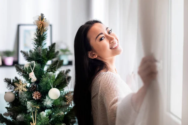Gelukkig aziatische vrouw staan in de buurt venster in kerst versierd huis — Stockfoto