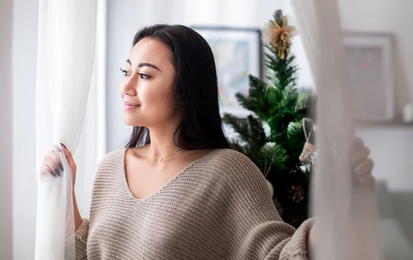 Glückliche Asiatin steht am Fenster in weihnachtlich dekoriertem Haus — Stockfoto