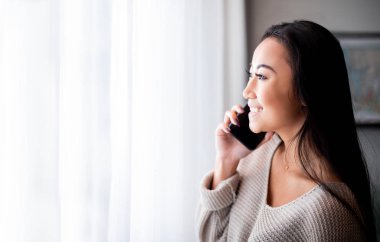 Akıllı telefon kullanan Asyalı bir kız ve pencerenin yanında duran bir evden arıyor.