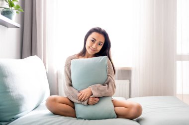 Evinde yastıkla kanepede oturan tatlı Asyalı genç bayan.