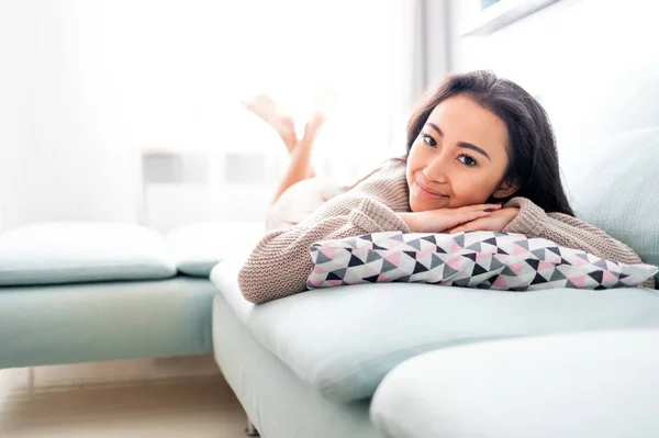 Šťastná asijská dívka ležící na pohovce s polštářem doma — Stock fotografie