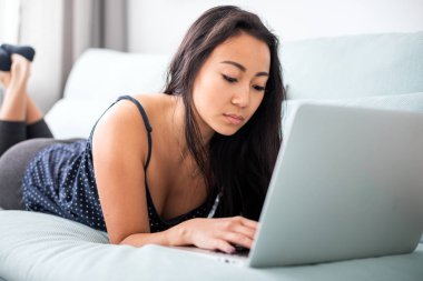 Evdeki kanepede dizüstü bilgisayar kullanan gülümseyen Asyalı genç kadın.