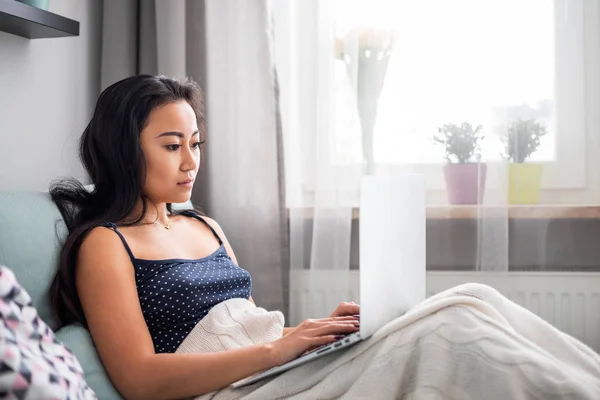 Розслаблена азіатська дівчина використовує ноутбук, сидячи на дивані вдома — стокове фото