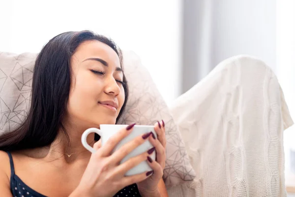 Avslappnad ung asiatisk kvinna sitter i fåtölj med filt dricka kaffe eller te — Stockfoto