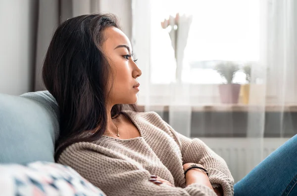 Sad asian girl sitting on sofa at home and thinking — Stock Photo, Image