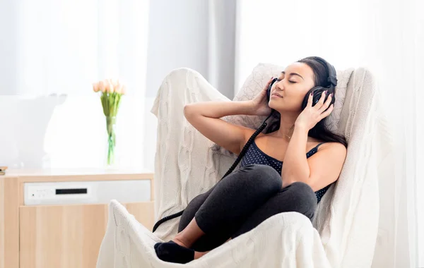 Sorrindo mulher asiática ouvindo música com fones de ouvido enquanto descansa em uma poltrona em casa — Fotografia de Stock