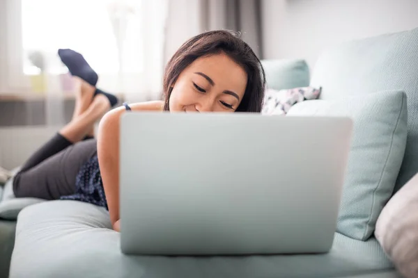 Sonriente asiático joven mujer usando portátil acostado en sofá en casa —  Fotos de Stock