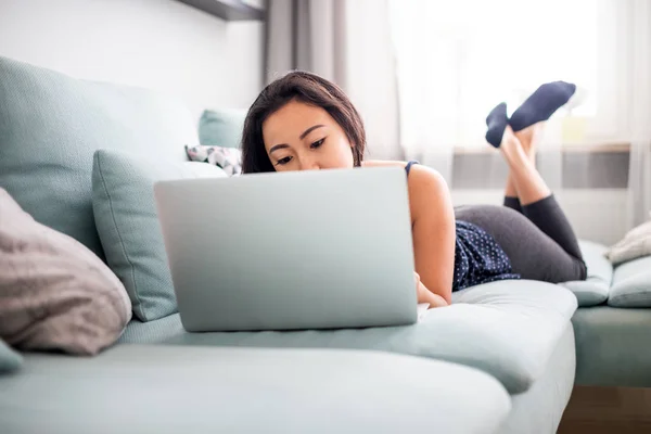 Усміхнена азіатська молода жінка за допомогою ноутбука лежить на дивані вдома — стокове фото